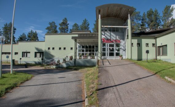 Hotellirakennuksen julkisivu. Keskellä katettu sisäänkäynti ja oven päällä nimi Tulikettu. Siitä vasemmalle laskeutuu portaikko ja yksikerroksinen rakennus laskeutuu kolmessa osassa kohti kuvan vasenta reunaa. Oikealla sisäänkäynnille johtava asfalttiluiska ja kuvaajaa kohti oikeassa reunassa suorakulmaisesti tuleva yksikerroksinen siipi. Rakennuksen takana on mäntyjä, etualalla asfalttipiha.